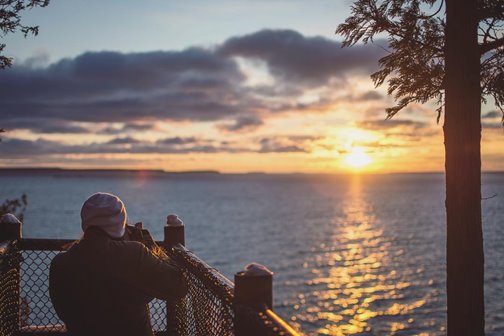 A woman bundled up taking a picture of the sunrise at the lake.