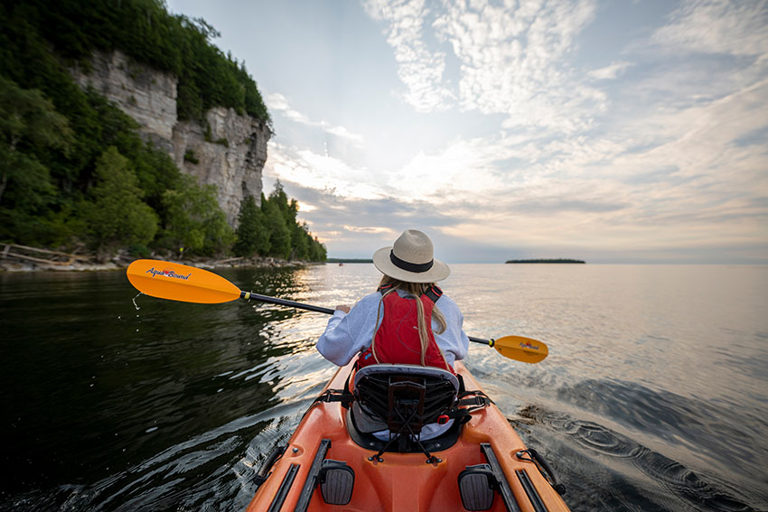 Point of view from the back of a kayak looking at the front person in still water by the cliffs