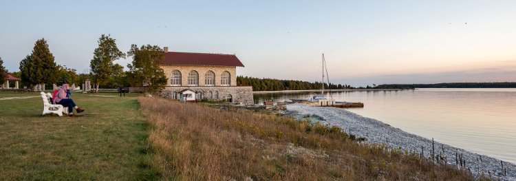 People sitting on a bench looking out at the sunsetting over the lake with the stone boathouse in the background