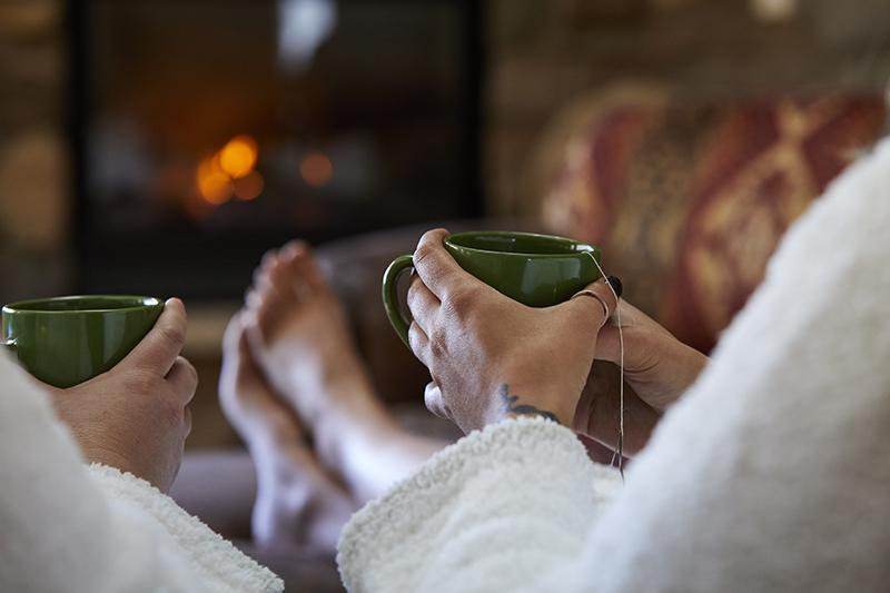 Two women relax in robes with mugs of tea in front a fireplace at spa