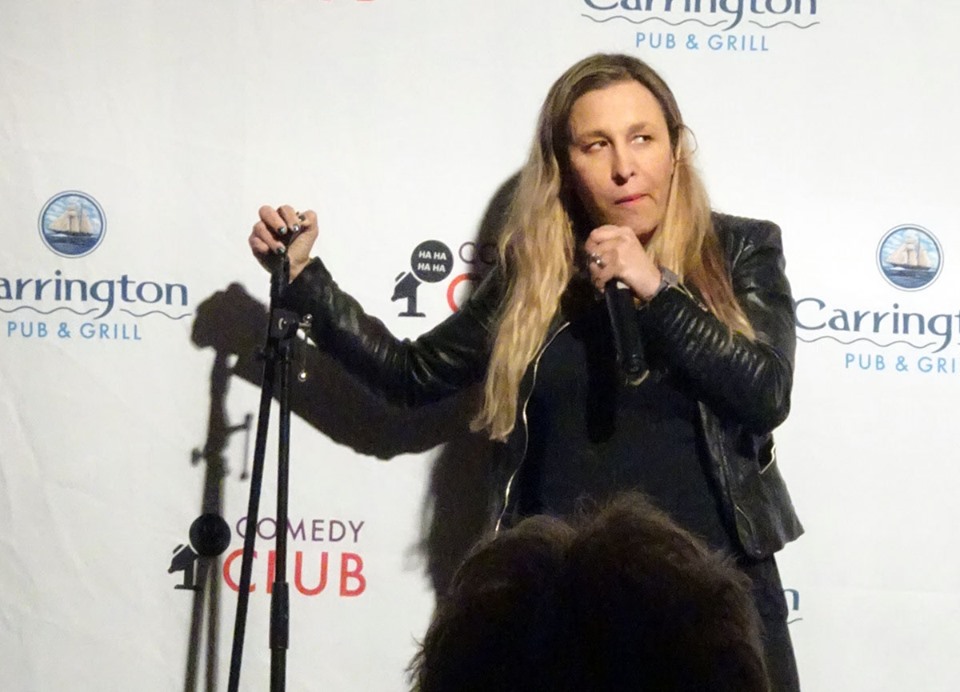 A standup comedian stands ion a stage in front of a microphone.