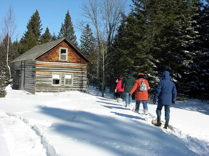 A family snowshoes near an old wooden cabin in the woods.