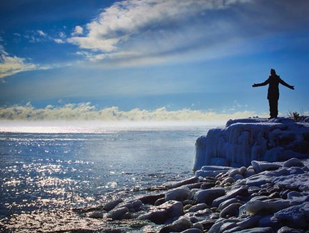 A person standing with their arms open looking out at the lake in winter