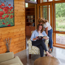 Woman kissing the top of another woman's head who is seated reading a book.