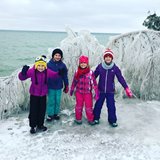 Children bundled up and smiling in front of ice-covered trees at the lakefront.