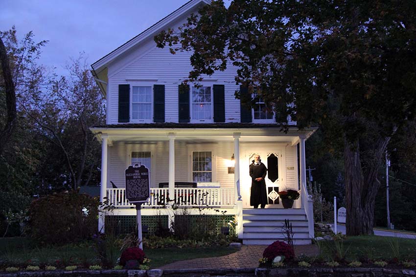 A creepy tour guide stands on the steps of the Alexander Noble House at dusk.