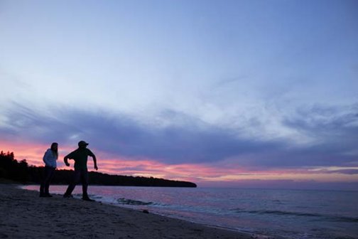 The silhouette of two people skipping rocks at the lakefront at sunrise.